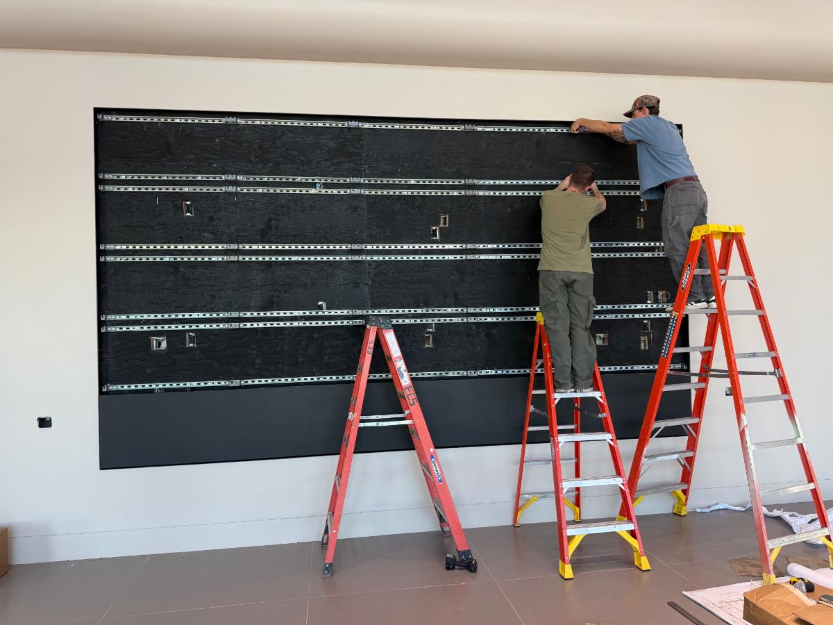 Installation visualization showing two installers on ladders aligning a StrutWall video wall frame.